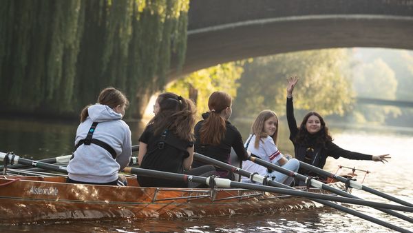 Fünf Schülerinnen und Schüler rudern in einem Boot unter einer Brücke hindurch.
