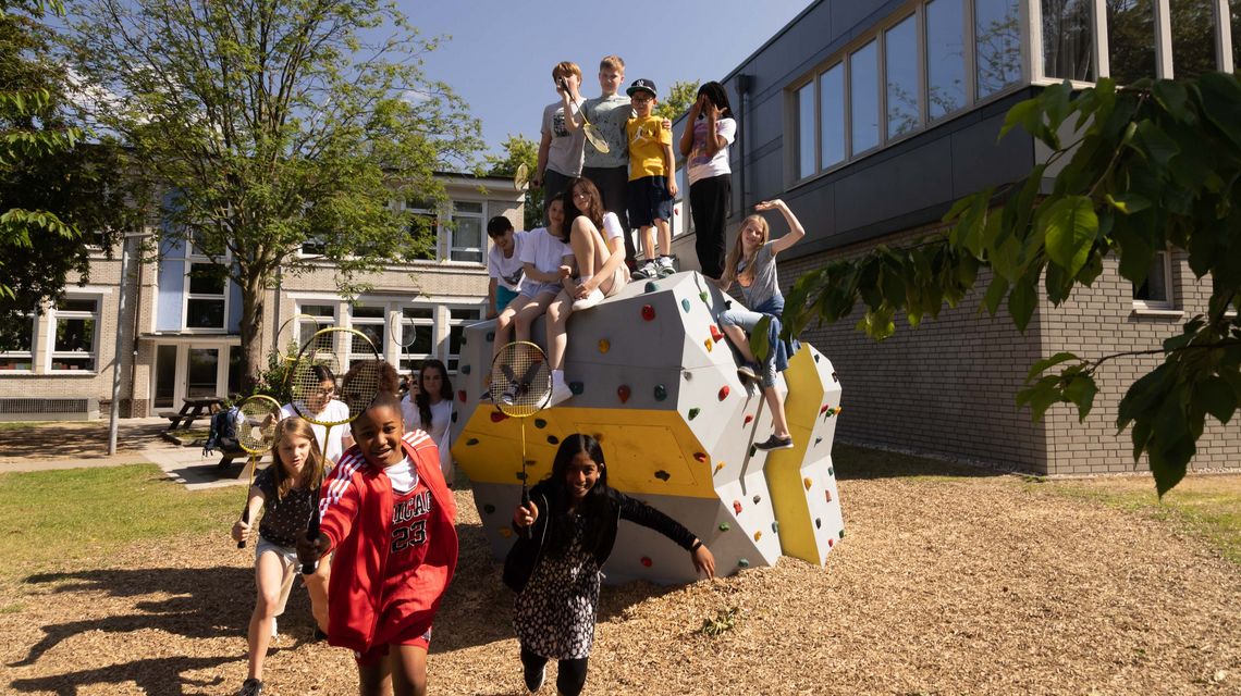 Eine Gruppe von Kindern steht oben auf einem Kletterfelsen auf dem Schulhof, einige lspielen Federball.