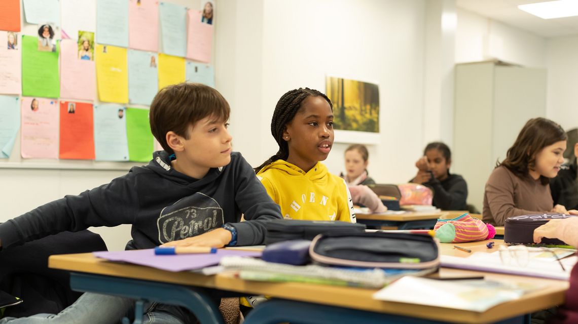 Blick in eine 5. Klasse im Unterricht. Im Vordergrund sitzen ein Mädchen in einem gelben Pullover und ein Junge.