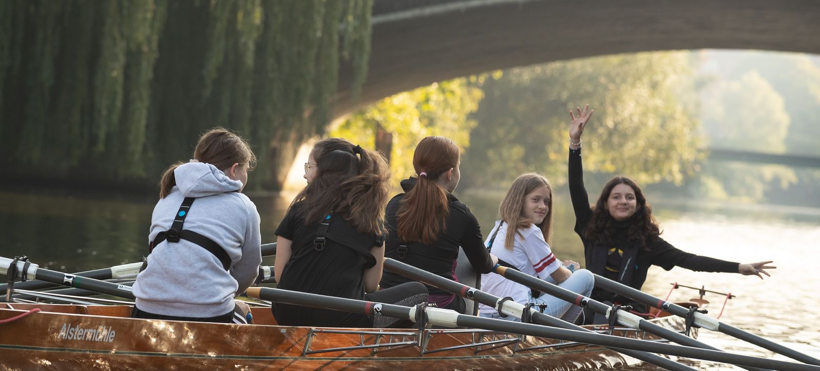 Fünf Schülerinnen und Schüler rudern in einem Boot unter einer Brücke hindurch.